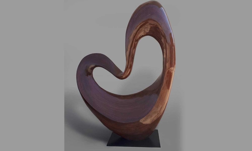 This image features a modern abstract wood sculpture. The design features a flowing, curved shape that elegantly twists and turns, creating a sense of movement and fluidity. The smooth, glossy finish of the wood highlights the elegant lines of the sculpture, while the warm, rich tones of the wood enhance its natural beauty. The sculpture is mounted on a simple black metal base, allowing the artwork to stand out and act as a focal point. Its unique, organic shape evokes a sense of balance and harmony, making it a stunning piece of modern.
