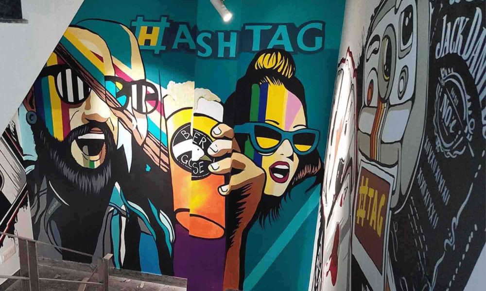 The walls of the stairwell are adorned with vibrant and colorful street art, in the form of stylized representations of a man and a woman, characterized by bold lines and intense colors. "Hashtag" fills the background with an image of a beer mug and sunglasses.